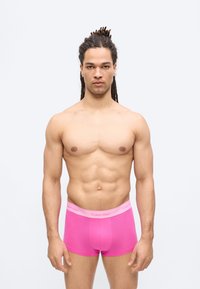 Μυώδης άνδρας με dreadlocks στέκεται προς τα εμπρός φορώντας φωτεινά ροζ εσώρουχα Calvin Klein, σε απλό ανοιχτό φόντο.