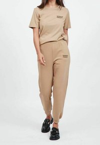 Beżowa bawełniana koszulka z krótkim rękawem oraz dopasowane dresy z elastycznym pasem; koszulka ma subtelny napis. Czarne lordsy dopełniają całość.