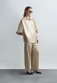 Cămașă bej din satin cu mâneci supradimensionate și guler, asortată cu pantaloni bej din satin cu picioare largi. Pantofi cu toc ascuțiți negri și ochelari de soare.