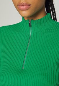 Haut zippé vert côtelé avec un col montant, doté d'une fermeture éclair métallique et d'un tissu texturé. Vue rapprochée mettant en valeur le design et la texture.