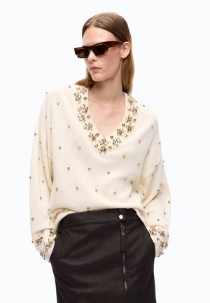 Donna con occhiali da sole tartarugati, maglione crema con decorazioni dorate e gonna nera a vita alta con zip frontale, mani nelle tasche.