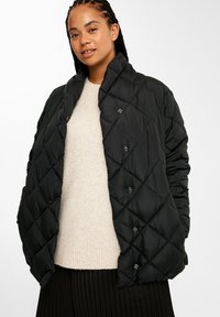 Schwarze gesteppte Pufferjacke mit Stehkragen, ausgestattet mit Knopfverschlüssen und einer glatten, glänzenden Textur. Über einem hellbeigen Pullover getragen.