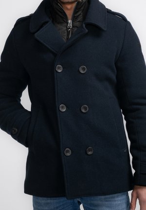 Hombre lleva un abrigo tipo pea coat de doble botonadura en azul marino oscuro con seis botones, con la mano en el bolsillo, sobre una chaqueta negra acolchada con cremallera y jeans azules.