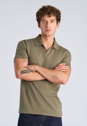 Joven con cabello rizado, vistiendo un polo de manga corta color verde oliva, con los brazos cruzados, mostrando tatuajes en ambos antebrazos, fondo neutro y claro.