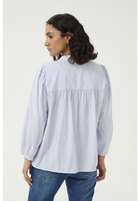 Blusa a righe blu chiaro con maniche lunghe a sbuffo, dettaglio arricciato sul retro e sottili puntini testurizzati. Indossata con jeans blu.