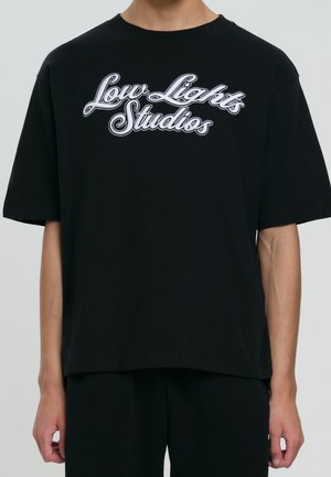 Osoba nosząca luźną czarną koszulkę z białym kursywowym napisem "Low Lights Studios" na piersi.