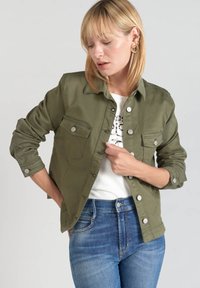 Giacca camicia verde oliva in cotone, con due tasche frontali a pattina, chiusura a bottoni e maniche arrotolate. Indossata sopra una maglietta bianca e jeans.