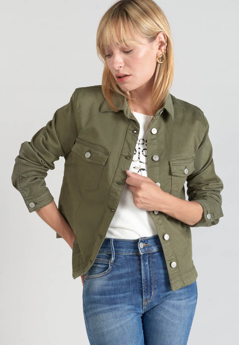 Giacca camicia verde oliva in cotone, con due tasche frontali a pattina, chiusura a bottoni e maniche arrotolate. Indossata sopra una maglietta bianca e jeans.