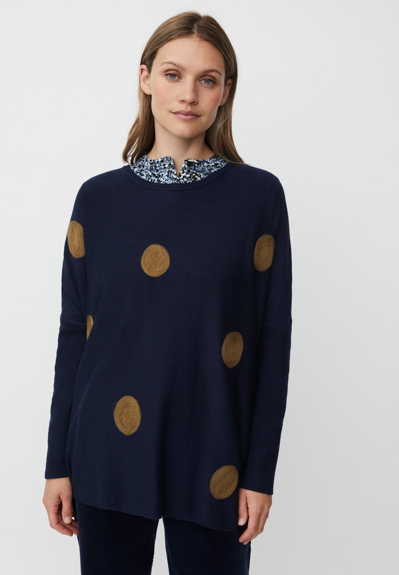 Granatowy sweter z dużymi brązowymi filcowymi kropkami, luźny krój, ribbowe mankiety oraz niebieska koszula w kwiatowy wzór z kołnierzykiem pod spodem.