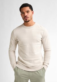 Light beige sweater with textured stripes, round neckline, and ribbed cuffs. Paired with olive green shorts, featuring side pockets.