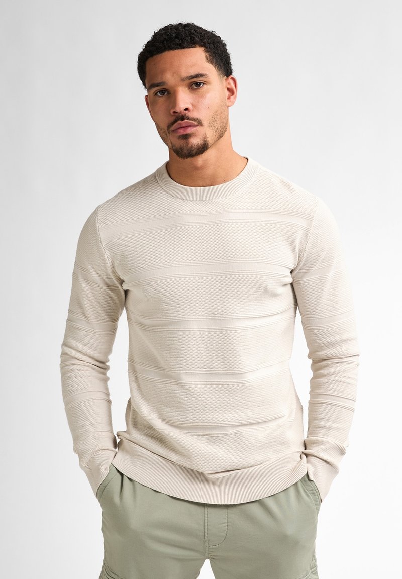 Light beige sweater with textured stripes, round neckline, and ribbed cuffs. Paired with olive green shorts, featuring side pockets.