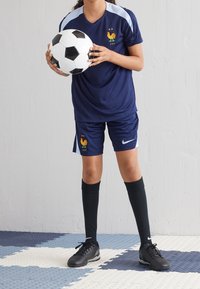 Námořnický fotbalový dres s jemně modrými akcenty, zdobený emblémem kohouta, doplněný černými ponožkami a kopačkami, který drží černobílý fotbal.