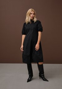 Šviesiaplaukė moteris dėvi juodą kelių ilgio suknelę su pūstomis rankovėmis ir juodus batelius su aukštais kulnais ir smailiais priekiniais galiukais, stovinti prie rudai pilkos spalvos fono.