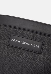 Borsa in pelle nera con superficie texturizzata e un'etichetta con marchio in lettere argentate che recita "TOMMY HILFIGER". Bordo liscio, design minimal.