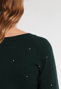 Épaule et haut du dos d'une personne aux cheveux châtain clair portant un pull vert foncé orné de petits clous dorés sur fond blanc.