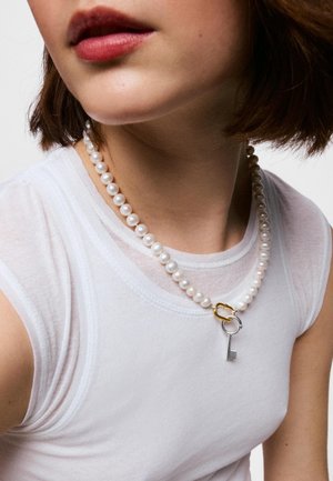 Femme portant un haut blanc sans manches et un collier de perles avec un pendentif clé en or et argent près du cou.