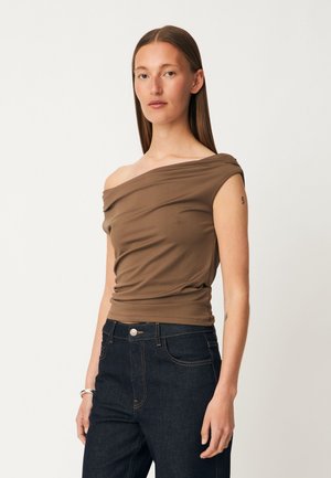 Kvinde med glat brunt hår iført en asymmetrisk brun top og mørkeblå jeans mod en ensfarvet hvid baggrund.