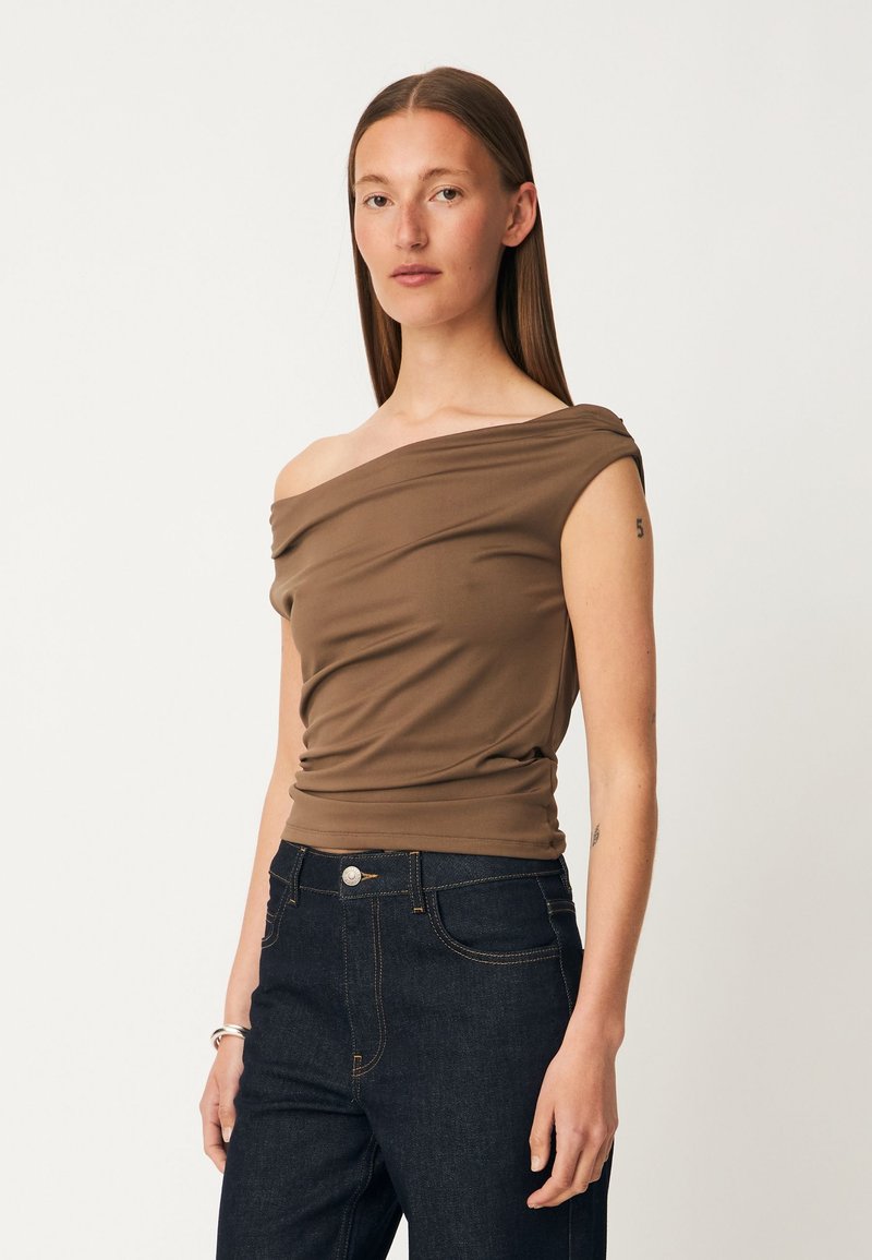 Femme aux cheveux bruns raides portant un haut marron asymétrique et un jean bleu foncé sur un fond blanc uni.