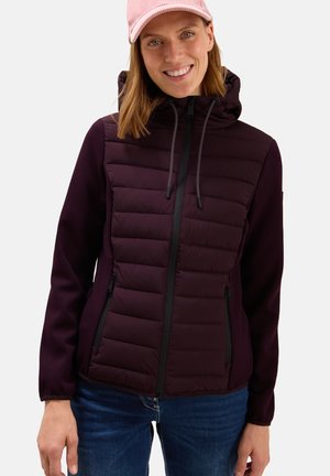 Smiling woman wearing a quilted dark purple hooded jacket, pink cap, and blue jeans, standing against a plain light background.