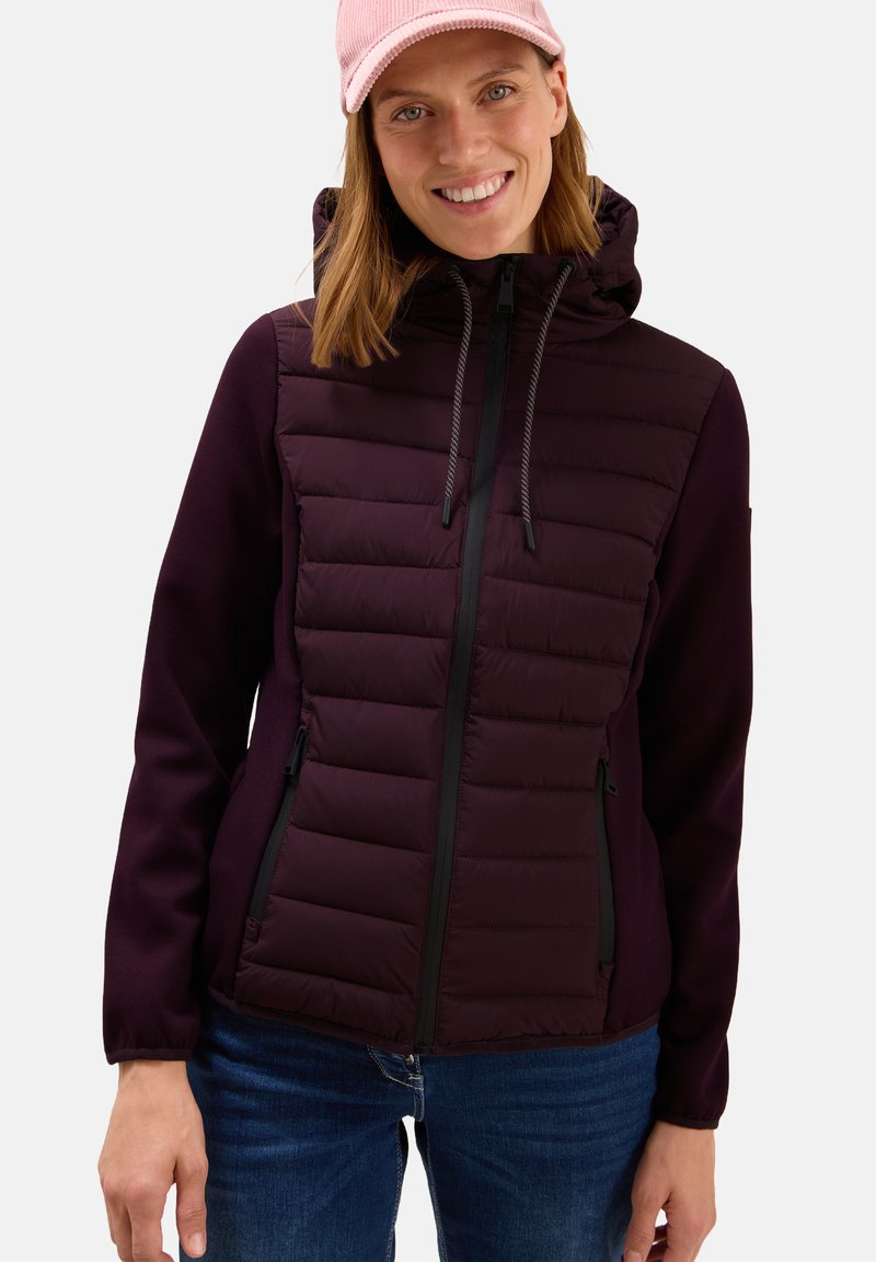 Femme souriante portant une veste à capuche matelassée de couleur violet foncé, une casquette rose et un jean bleu, se tenant devant un fond clair.