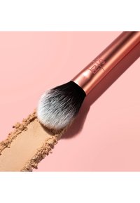 Pinceau à maquillage avec des poils noirs et blancs balayant une poudre libre beige sur une surface rose, montrant un manche en or rose marqué "402".