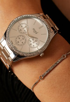 Montre-bracelet en argent avec lunette ornée de cristaux et trois sous-cadrans au poignet, associée à un délicat bracelet en cristal sur une peau bronzée.