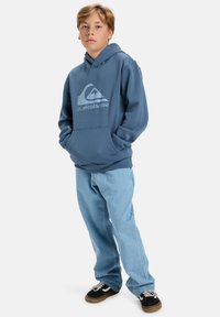 Blue hooded sweatshirt with a front pocket and logo, paired with light blue jeans and black sneakers with white laces.