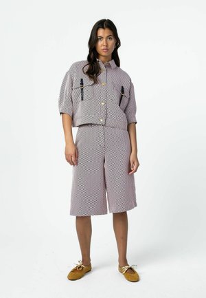 Kobieta w dopasowanej, wzorzystej koszuli z krótkim rękawem z klamrami oraz szerokich, sięgających do kolan szortach, do tego brązowe baleriny z kokardkami.