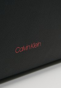 Surface texturé noir avec le logo "Calvin Klein" en rouge. Le matériau semble être du simili cuir. Design simple et épuré.