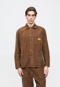 Ung mann med kort brunt hår iført brun kordfløyelsjakke og matchende bukser, stående mot en enkel hvit bakgrunn.