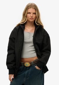 Sort zip-up bomberjakke med hætte, parret med en grå ribbet crop top og højtsiddende blå denim jeans. Bemærkelsesværdig guld bæltet spænde som accent.