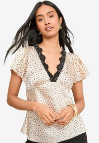 Blusa color crema con pois neri, con merletto lungo il collo a V e maniche a sbuffo. La trama morbida e simile al satin aggiunge eleganza.