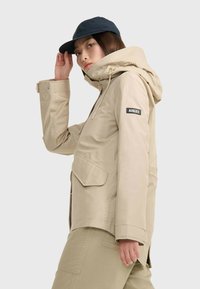 Beige wasserdichte Jacke mit Kapuze, mit Fronttaschen und verstellbaren Bündchen. Ein schwarzes Logo-Patch befindet sich am linken Ärmel.