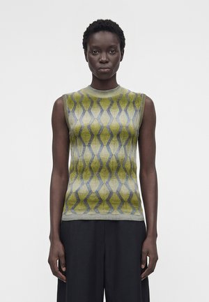 Femme portant un haut en maille sans manches à motifs verts et gris avec un pantalon noir à jambes larges, se tenant devant un fond clair uni.