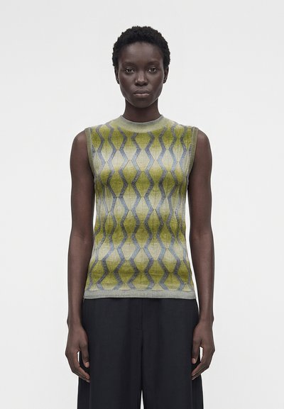 Femme portant un haut en maille sans manches à motifs verts et gris avec un pantalon noir à jambes larges, se tenant devant un fond clair uni.