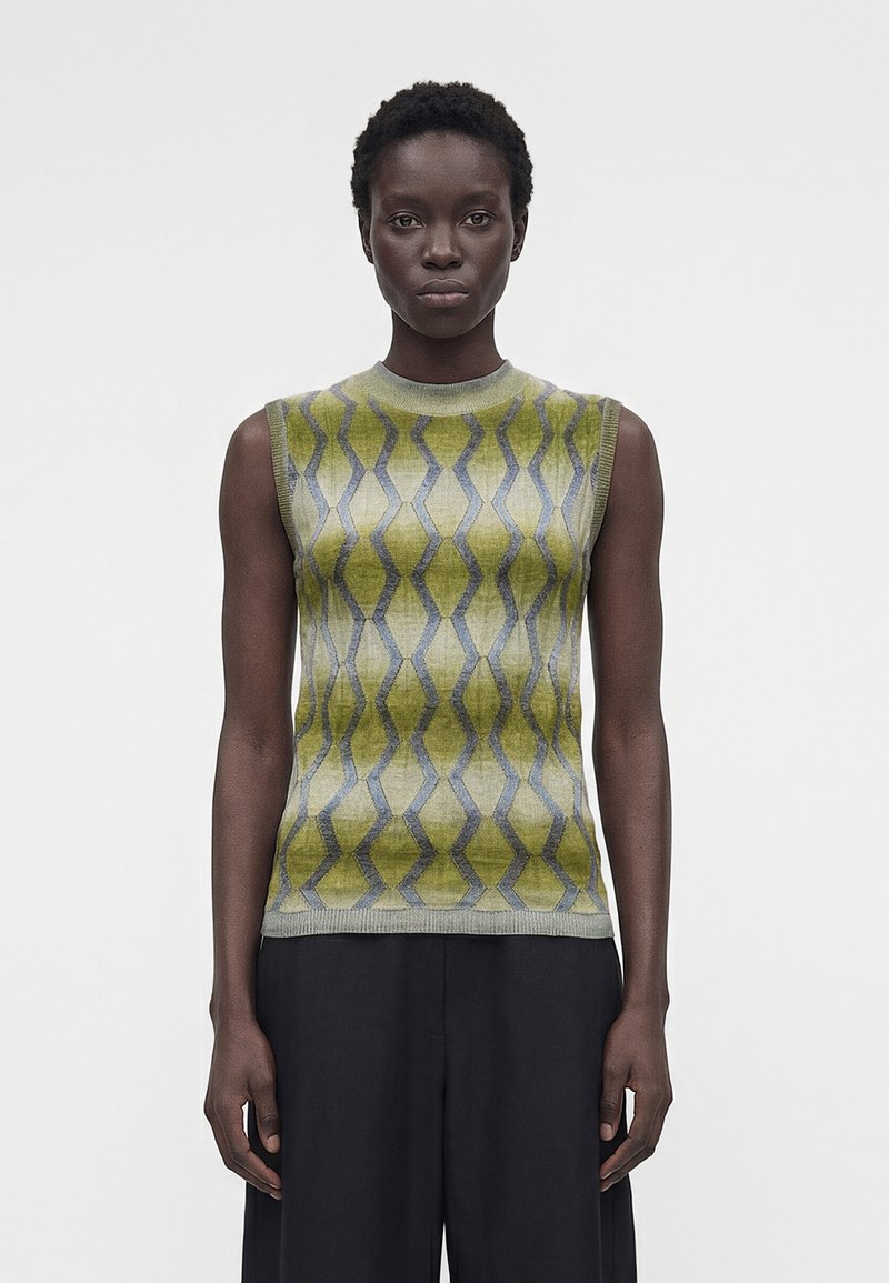 Γυναίκα φοράει αμάνικο πλεκτό τοπ με πράσινο και γκρι σχέδιο και μαύρο παντελόνι με φαρδιά μπατζάκια, στέκεται μπροστά σε απλό φωτεινό φόντο.