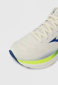Chaussure de sport beige clair avec une tige texturée, des accents bleus et vert fluorescent, et un bout arrondi. Dispose de lacets plats et d'une semelle rembourrée.