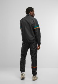 Chándal deportivo negro con franjas en contraste de color teal y naranja, con un corte relajado y puños elásticos. Modelo visto desde atrás.