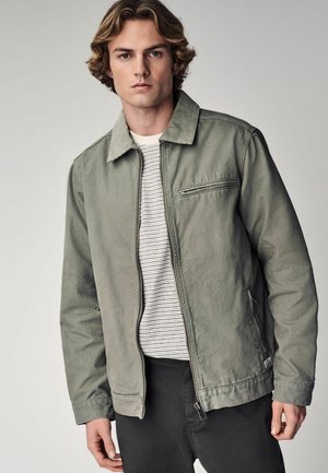 Chaqueta de algodón color verde oliva con cierre de cremallera, cuello con punta y un bolsillo en el pecho. Usada sobre una camisa a rayas, con un ajuste relajado.
