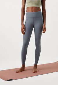 Højtaljede grå leggings lavet af strækstof med sidesømme. Båret med en lys tanktop og står på en lyserød yogamatte.