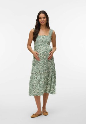 Zwangere vrouw die staat, gekleed in een mouwloze midi-jurk met een groen-wit patroon en bruine platte schoenen, met haar handen rustend op haar buik.