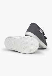 Zapatillas de lona grises con suelas de goma blancas y correas de Velcro. La parte superior cuenta con un tejido texturizado con un diseño cosido y una etiqueta de marca.