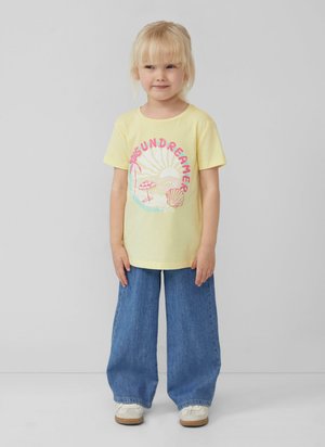 Gelbes T-Shirt mit "SUN DREAMER" Grafik; kombiniert mit lockeren, blauen Jeans. Kind trägt weiße Schuhe. Einfarbiger Hintergrund.