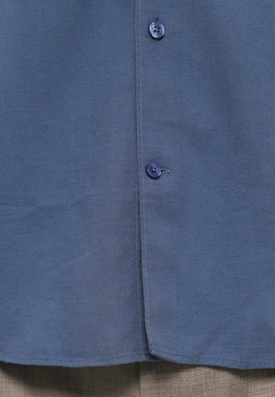 Close-up van blauwe gebuttonde overhemdstof met twee zichtbare knopen over beige textuurbroek.
