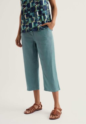 Persoon draagt teal wijde cropped broek, blauwgroen patroon top en bruine leren sandaaltjes met bandjes, staat met één hand in de zak.