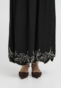 Robe noire longueur sol avec broderie de feuilles blanches à l'ourlet. Présente une texture douce et un ourlet en forme de vagues. Chaussures brunes à bout pointu visibles.