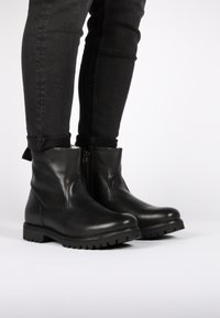 Botas de tobillo de cuero negro con un acabado suave, punta redonda y suela de goma texturizada; cuentan con un cierre lateral y costuras mínimas.