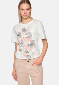 White short-sleeve t-shirt featuring black floral sketches and light pink text. Worn with light beige pants, showing a relaxed fit.
