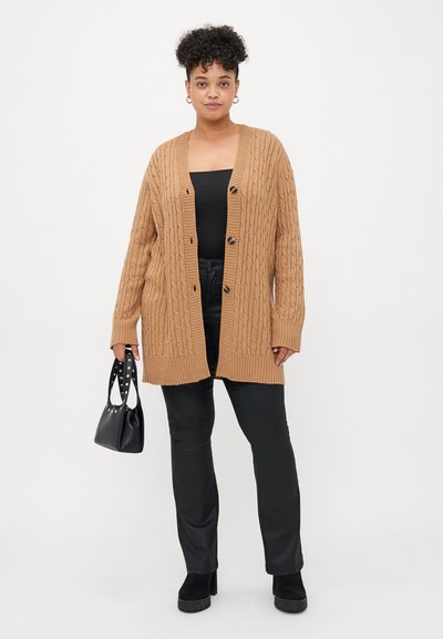 Jeune personne ayant les cheveux bouclés courts, portant un cardigan en tricot torsadé beige par-dessus un haut et un pantalon noirs, tenant un sac à main noir orné de clous en argent.
