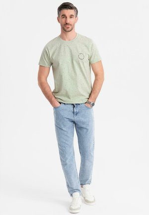 Camiseta de manga corta verde claro con motas negras y un logo en el pecho, combinada con jeans de corte relajado azul claro y zapatillas blancas.